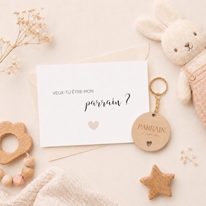 Peut inclure: Une carte avec le texte "VEUX-TU ÊTRE MON parrain ?" et un cœur, ainsi qu'un porte-clés en bois avec l'inscription "PARRAIN d'amour". Un lapin en peluche et une étoile en bois sont également présents. Les objets sont disposés sur une surface claire.