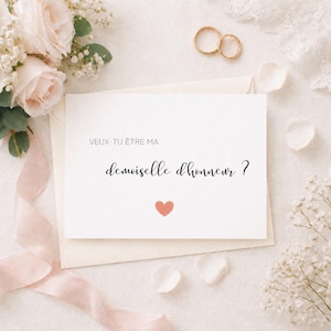 May include: A white card with the text "VEUX-TU ÊTRE MA demoiselle d'honneur ?" and a small coral heart. The card is on a cream envelope, surrounded by flowers, petals, and gold wedding rings, suggesting a wedding theme.