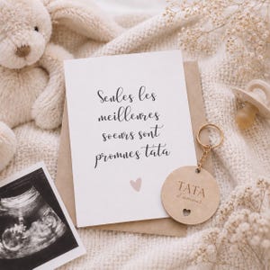 Peut inclure: Une carte d'annonce de bébé avec le texte "Seules les meilleures soeurs sont promues tata" et un petit cœur rose. Un porte-clés en bois avec le texte "TATA d'amour" et une découpe en forme de cœur. Une image d'échographie et un ours en peluche beige.
