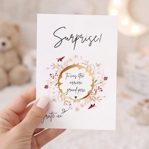 May include: A white rectangular card featuring the word Surprise! in black script at the top. Below, a circular gold scratch-off area is surrounded by a delicate floral wreath. The text Gratte ici with an arrow points to the gold circle.
