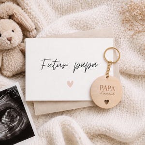 May include: A key chain with a wooden round pendant that says "PAPA d'amour" with a heart cutout, and a card that says "Futur papa" with a pink heart. A stuffed bunny and an ultrasound image are also in the image.