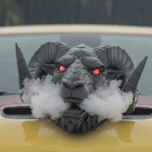 以下が含まれることがあります： 黄色い車のボンネットに取り付けられた、赤い目が光る暗い灰色の雄羊の頭の彫刻。彫刻は口から白い蒸気を出し、劇的な効果を生み出しています。雄羊は大きく湾曲した角と詳細な顔の特徴を持っています。