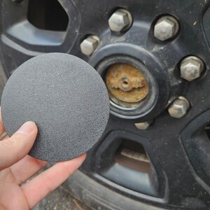 May include: A dark gray, textured hub cap held in front of a black wheel. The wheel has silver lug nuts and a rusty center. The hub cap is round and appears to be made of plastic.