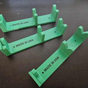 May include: Three green plastic display stands. Each stand has a curved base with two upright supports and a central support. The text "MADE IN USA" is printed on each stand.