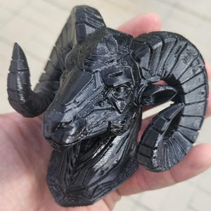 May include: A black, 3D-printed ram's head sculpture. The sculpture has detailed textures and curved horns. The ram's head is held in a hand, showing the size and detail of the artwork. The sculpture is a decorative item.