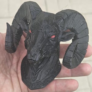 Könnte beinhalten: Eine schwarze Widderkopfskulptur mit roten Augen. Der Widder hat große, gebogene Hörner und eine strukturierte Oberfläche. Die Skulptur wird in einer Hand gehalten, die die Details des 3D-Druckdesigns zeigt.