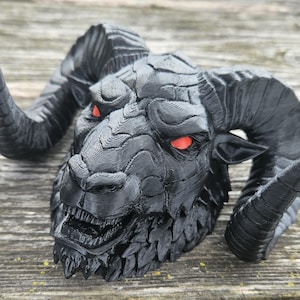 May include: A black ram's head sculpture with large, curved horns and red eyes. The sculpture has a detailed, textured surface and an open mouth, giving it a menacing appearance. The ram's head is a decorative item.