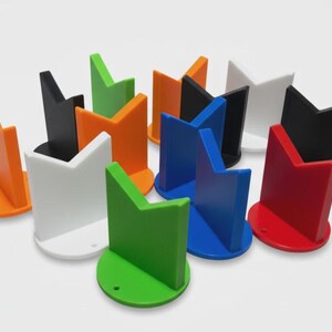 May include: A collection of colorful, geometric phone stands in various colors, including orange, black, white, green, blue, and red. Each stand has a circular base and a stylized, angled top, designed to hold a phone upright.