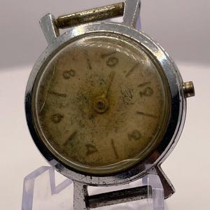 May include: A vintage wristwatch with a silver metal case and a worn, gold-toned face. The watch has black numerals and hands, and is displayed on a clear plastic stand. The watch appears to be an antique.