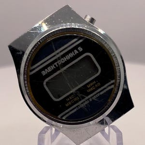 Puede incluir: Un reloj de pulsera digital vintage con esfera negra y caja plateada. La esfera del reloj muestra el texto "Электроника 5" y otros caracteres cirílicos. El reloj se exhibe sobre un soporte de plástico transparente.