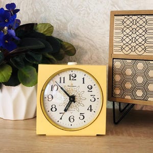 Könnte beinhalten: Ein quadratischer, gelber Vintage-Wecker mit weißem Zifferblatt und goldfarbenem Rand. Das Zifferblatt hat schwarze Ziffern und Zeiger mit einem dekorativen Blumendesign in der Mitte. Die Uhr ist etwa 10 cm breit.