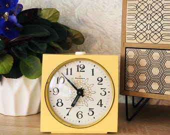 May include: A square, yellow vintage alarm clock with a white face and gold trim. The clock face features black numerals and hands, with a decorative floral design in the center. The clock is approximately 4 inches wide.