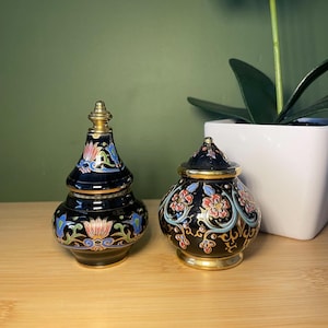 Può includere: Due vasi decorativi neri con coperchi. I vasi hanno dettagli dorati e sono adornati con motivi floreali colorati in rosso, blu e rosa. I vasi sono su una superficie di legno.