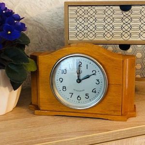 Op de afbeelding: Een houten mantelklok met een lichtbruine afwerking. De wijzerplaat is wit met zwarte cijfers en wijzers. De klok is rechthoekig met een gebogen bovenkant en staat op een houten oppervlak. Een kleine plant met paarse bloemen is zichtbaar.