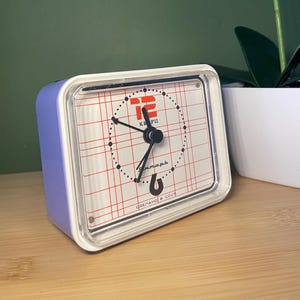 Peut inclure: Un réveil blanc et bleu avec un cadran rouge et noir. L'horloge a un motif de grille et le texte "Сделано в СССР" en bas. L'horloge est réglée sur 12 heures.