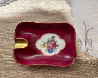 May include: A rectangular, burgundy ashtray with a gold rim and a floral design. The ashtray features a white, cloud-shaped centerpiece with a bouquet of pink, purple, and yellow flowers. The ashtray has a curved indentation for resting cigarettes.