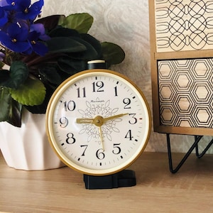 Può includere: Una sveglia vintage dorata con quadrante color crema, numeri neri e lancette dorate. L'orologio ha una base nera e un motivo floreale centrale. Sullo sfondo, una pianta in vaso con fiori viola. Il diametro è di circa 10 cm.