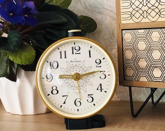 May include: A vintage gold-toned alarm clock with a cream-colored face, black numerals, and gold hands. The clock has a black base and a decorative floral design in the center. A potted plant with purple flowers is in the background.