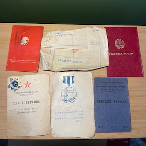 May include: A collection of vintage documents and ephemera, including red and blue booklets, a white envelope, and a medal. The items feature Cyrillic text and imagery, suggesting a historical or commemorative theme.
