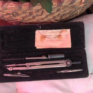 Puede incluir: Una caja de cuero negro con una brújula y una regla en su interior. La caja tiene un papel rosa con texto en ruso en su interior. El texto dice "приспособление для выполнения технических учебных работ с 2003 года."