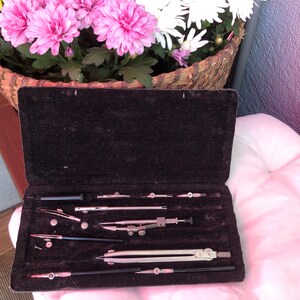 Può includere: Un set di compassi vintage in una custodia di velluto nero. Il set comprende un compasso, un divisore e un righello. Gli strumenti sono in metallo e hanno una finitura argentata.