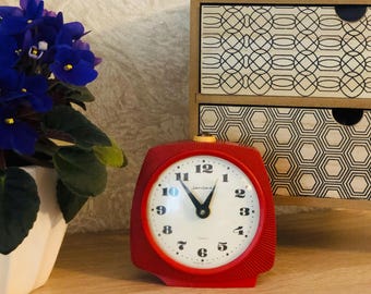 May include: A red vintage quartz clock with a white face and black numerals. The clock has a rectangular shape with rounded corners and a gold-colored top. A potted plant with blue flowers is to the left, and a small wooden cabinet is in the background.
