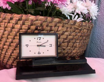 May include: A vintage black desk clock with a rectangular face, featuring a white background and black numerals. The clock is set on a tiered base with three small gold-colored rings. A woven basket filled with pink and white flowers is in the background.