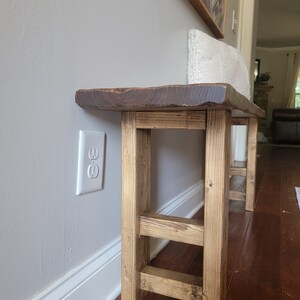 May include: A rustic wooden bench with a natural wood finish and a live edge top. The bench has a simple, farmhouse style and is supported by four legs with a cross brace.