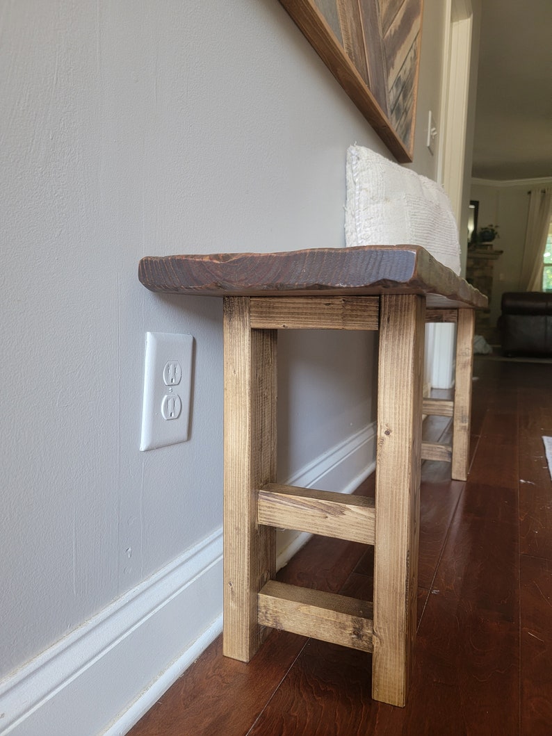 May include: A rustic wooden bench with a natural wood finish and a live edge top. The bench has a simple, farmhouse style and is supported by four legs with a cross brace.