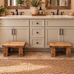 May include: Two low, rectangular wooden step stools with a natural, distressed finish and four sturdy legs, positioned on a tiled floor in front of a light gray bathroom vanity cabinet featuring dark metal hardware and a woven textured rug.