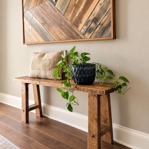 May include: A rectangular wooden bench with four legs, a beige textured rectangular pillow, and a dark blue patterned ceramic pot containing a green trailing plant. Above the bench hangs a large wooden wall art piece with a chevron pattern.