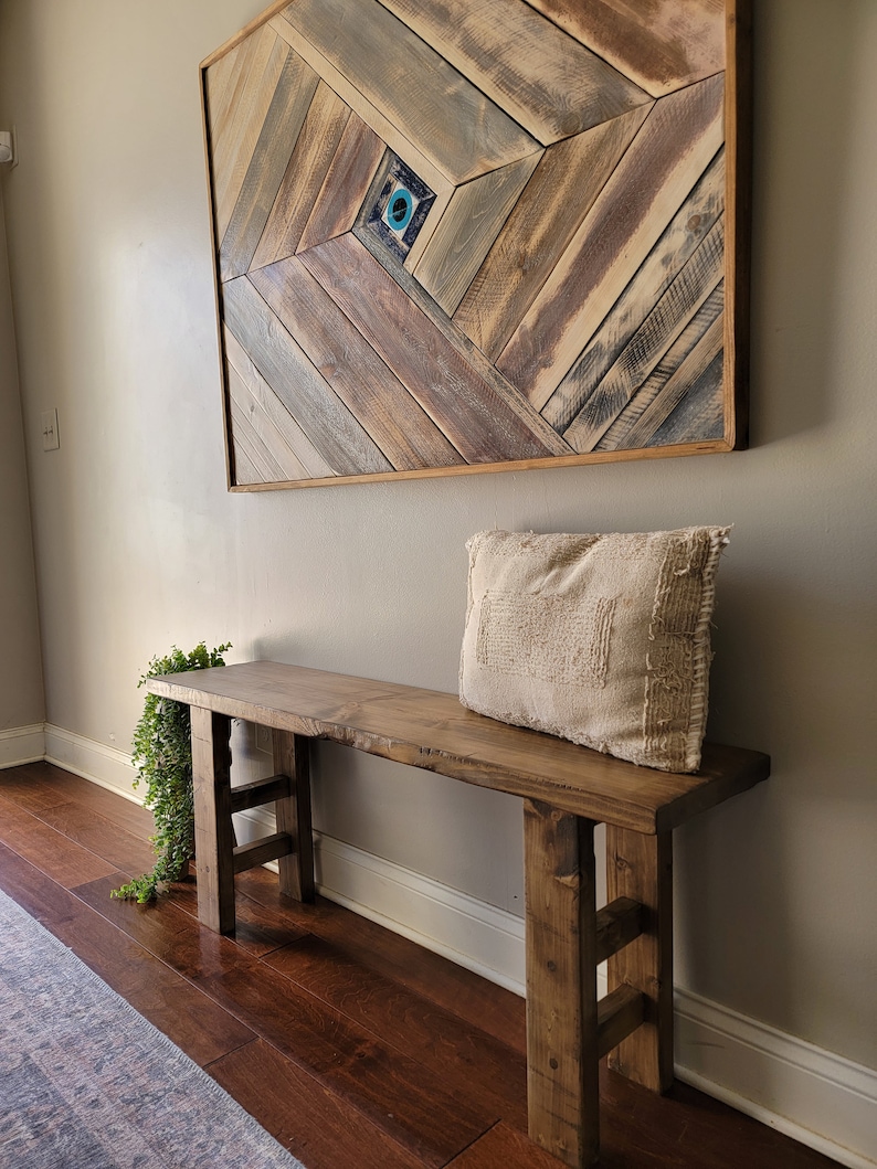 May include: A rustic wooden bench with a natural wood finish and a simple design. The bench has two legs on each side and a wide, flat top. A white throw pillow is placed on the bench. A wooden wall art piece with a geometric pattern is hanging on the wall behind the bench.