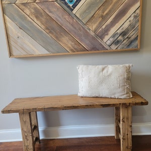 May include: A rustic wooden bench with a natural wood finish and a white cushion on top. The bench has two legs on each side and is made of reclaimed wood. The bench is in front of a wall with a wooden wall art piece featuring a blue and white eye design.