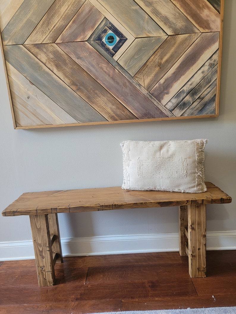 May include: A rustic wooden bench with a natural wood finish and a white cushion on top. The bench has two legs on each side and is made of reclaimed wood. The bench is in front of a wall with a wooden wall art piece featuring a blue and white eye design.