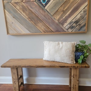 May include: A wooden bench with a natural finish and a white throw pillow. The bench has a simple design with two legs on each side. The bench is in front of a wall with a wooden wall art piece featuring a blue and white eye design.