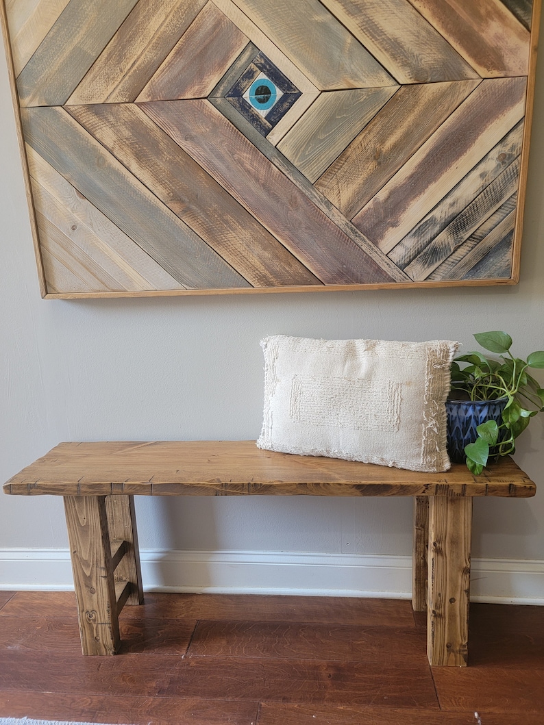May include: A wooden bench with a natural finish and a white throw pillow. The bench has a simple design with two legs on each side. The bench is in front of a wall with a wooden wall art piece featuring a blue and white eye design.