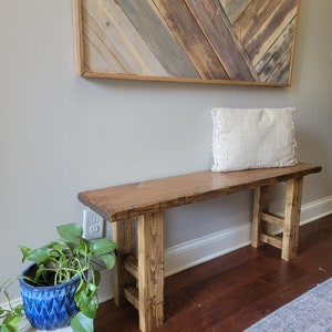 May include: A rustic wooden bench with a natural wood finish and a simple design. The bench has a wide, flat top and two sturdy legs. The bench is sitting on a wooden floor with a rug in the foreground.