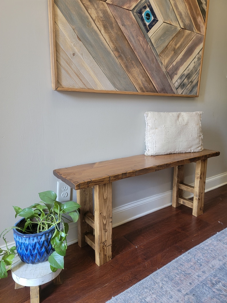May include: A rustic wooden bench with a natural wood finish and a simple design. The bench has a wide, flat top and two sturdy legs. The bench is sitting on a wooden floor with a rug in the foreground.