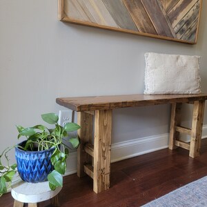 May include: A rustic wooden bench with a natural wood finish and a white throw pillow. The bench is sitting on a hardwood floor with a light blue rug in the foreground. A potted plant with blue and white patterned ceramic pot is on a white pedestal to the left of the bench.