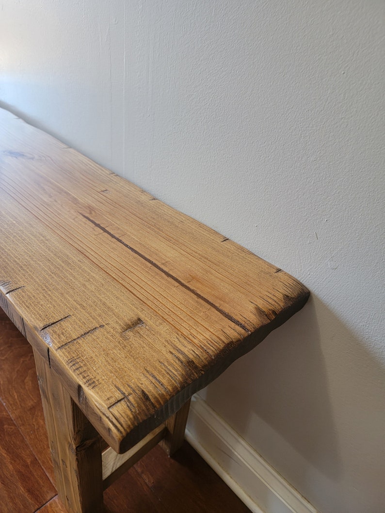 May include: A rustic wooden bench with a natural wood finish. The bench has a simple design with a wide, flat top and two legs. The wood has a natural grain pattern and is slightly distressed.