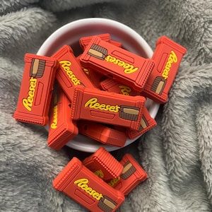 May include: A collection of red Reese's candy bar charms with yellow lettering and brown chocolate detailing. The charms are piled in a white bowl, set against a soft, gray background.