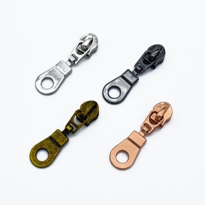 Op de afbeelding: Vier ritsluitingen in verschillende afwerkingen: zilver, zwart, goud en rosé goud. Elke ritsluiting heeft een lus aan de onderkant en een schuifregelaar in het midden.