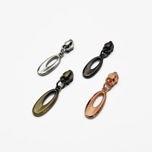 Könnte beinhalten: Vier Reißverschluss-Zipper in verschiedenen Farben: Silber, Schwarz, Gold und Roségold. Jeder Zipper hat einen ovalen Anhänger mit einem Loch in der Mitte.