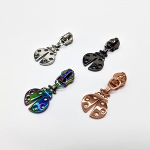 Könnte beinhalten: Vier Reißverschluss-Zipper in Form von Marienkäfern. Die Zipper sind in Silber, Schwarz, Regenbogen und Roségold.