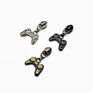 Op de afbeelding: Drie metalen ritstrekkers in zilver, zwart en brons, elk in de vorm van een videogamecontroller. De trekkers staan op een witte achtergrond, met de controllers iets schuin. De controllers hebben gedetailleerde knop- en richtingspadontwerpen.