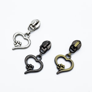 Può includere: Tre tiretti per cerniera con ciondoli a forma di cuore che presentano un'impronta di zampa. I tiretti per cerniera sono argentati, neri e dorati.