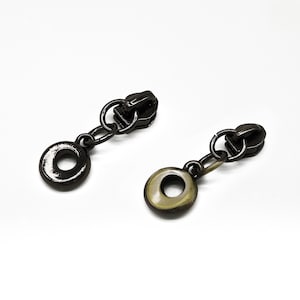 May include: Two black metal zipper pulls with a ring and a round charm attached to each. The charms are different colors, one is black and the other is gold.