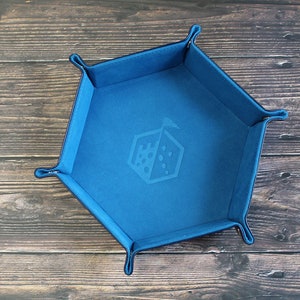 May include: A blue, hexagonal dice tray with a dice graphic in the center. The tray has a soft, felt-like texture and is designed to contain dice during tabletop gaming. The tray is shown on a wooden surface.