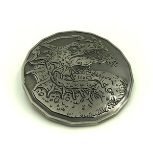 Puede incluir: Una moneda de metal plateada con un grabado detallado de una cabeza de dragón. El dragón tiene una expresión feroz con dientes afilados y un cuerpo largo y sinuoso.