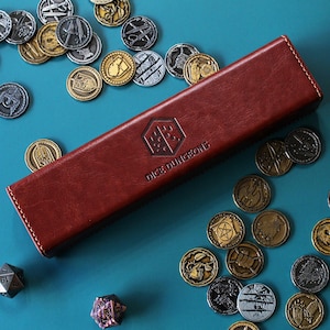 May include: A brown leather dice box with the text "Dice Dungeons" embossed on the top. The box is surrounded by a variety of metal dice with different designs.